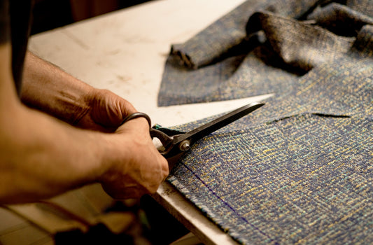Hands using large tailor's scissors to cut textured tweed fabric on work surface, photographed in closeup