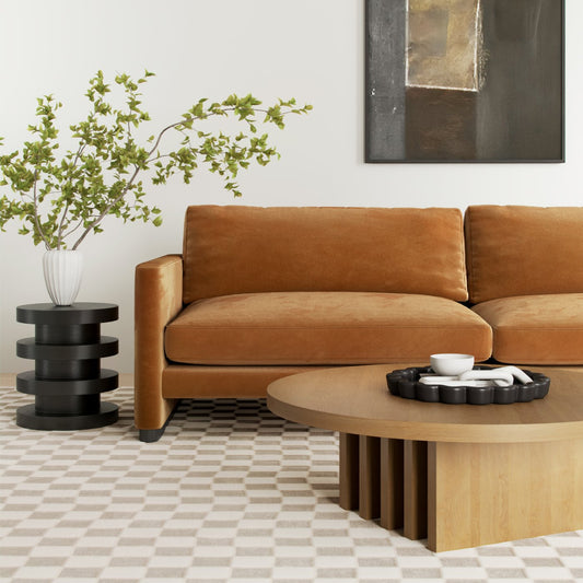Modern brown velvet sofa with round wood coffee table, black side table, and leafy plant in white vase
