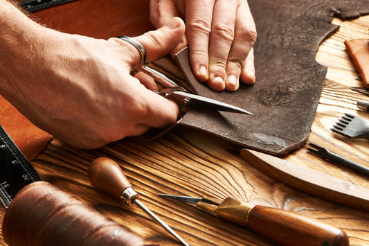 Craftsman cuts leather with scissors on wooden workbench with traditional leatherworking tools visible
