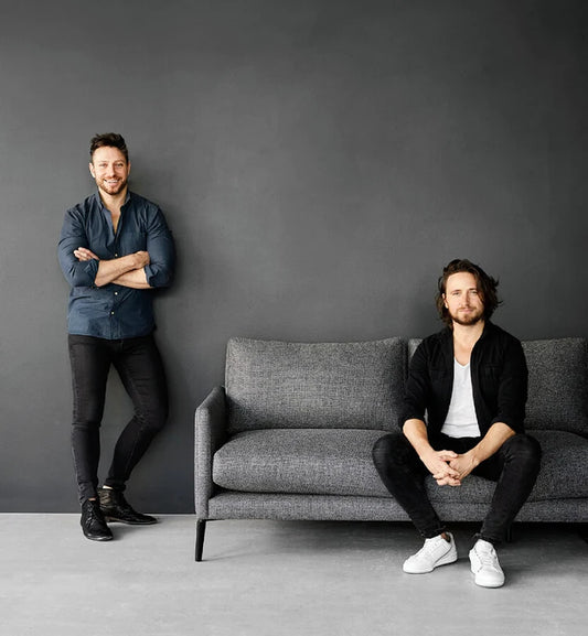 Two men in casual attire pose against grey wall, one standing, one seated on modern grey couch