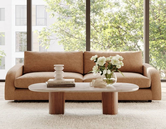 Modern camel sofa with oval marble coffee table, white flowers in vase, books and decor, bright window view