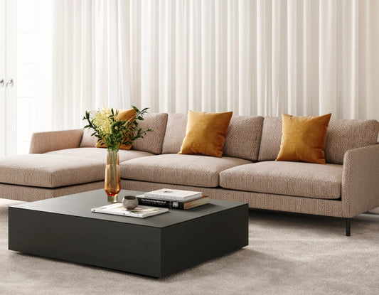 Modern L-shaped beige sectional with mustard pillows, dark coffee table, yellow flowers, white curtains in bright room